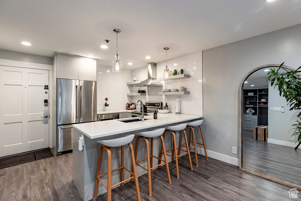 Kitchen featuring appliances with stainless steel finishes, wall chimney exhaust hood, a peninsula, dark wood-type flooring, and recessed lighting