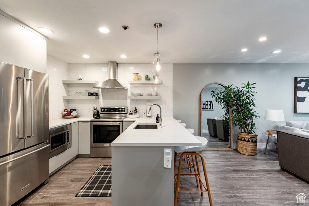 Kitchen featuring open shelves, appliances with stainless steel finishes, wall chimney range hood, a kitchen bar, and a peninsula