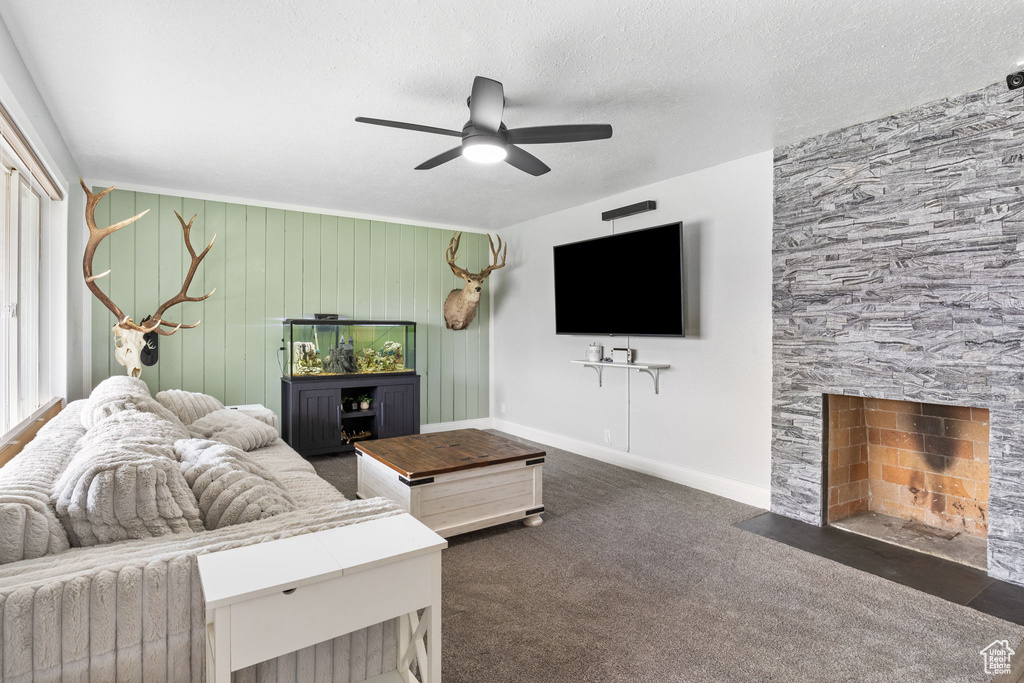 Living room with a ceiling fan, a stone fireplace, dark colored carpet, and a textured ceiling