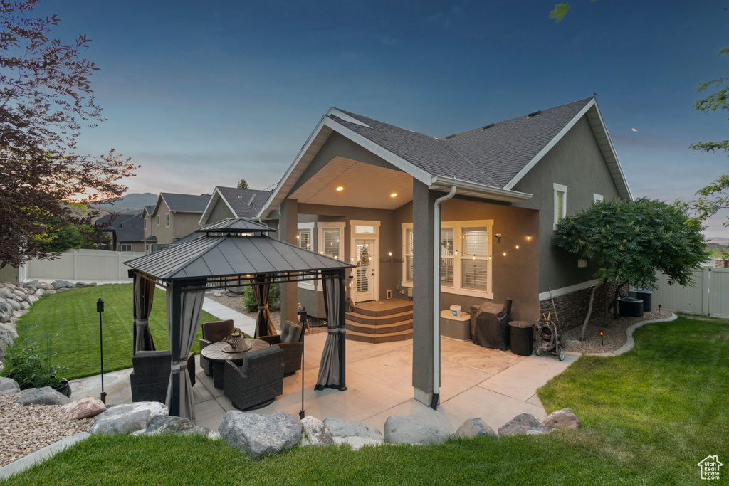 Back of house at dusk featuring stucco siding, a gazebo, a fenced backyard, a patio, and a shingled roof