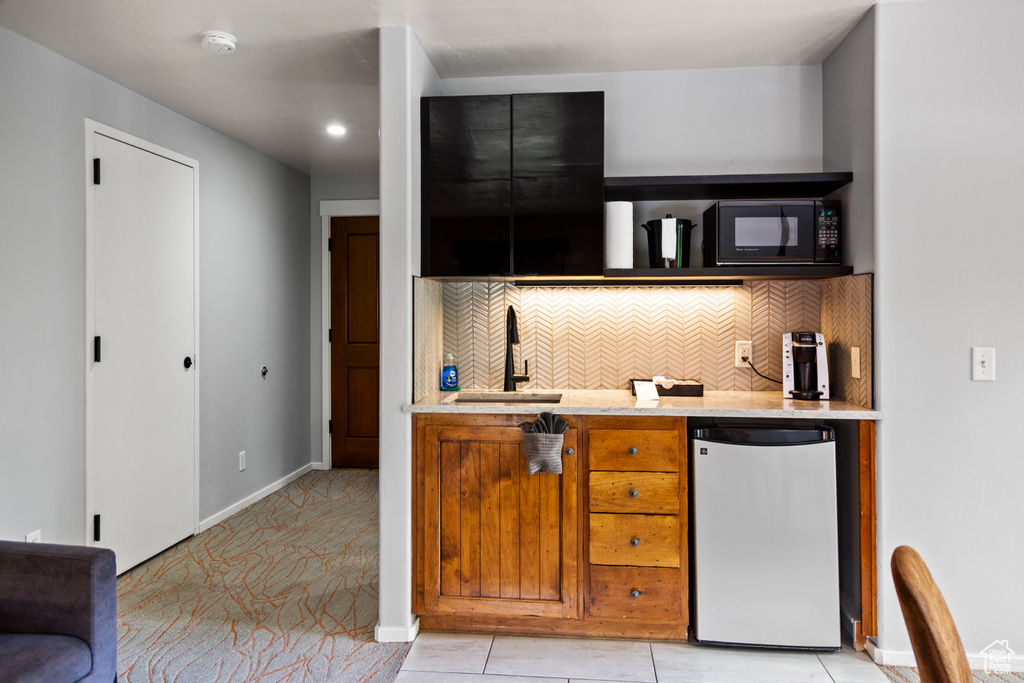Kitchen with refrigerator, black microwave, backsplash, light stone counters, and brown cabinetry