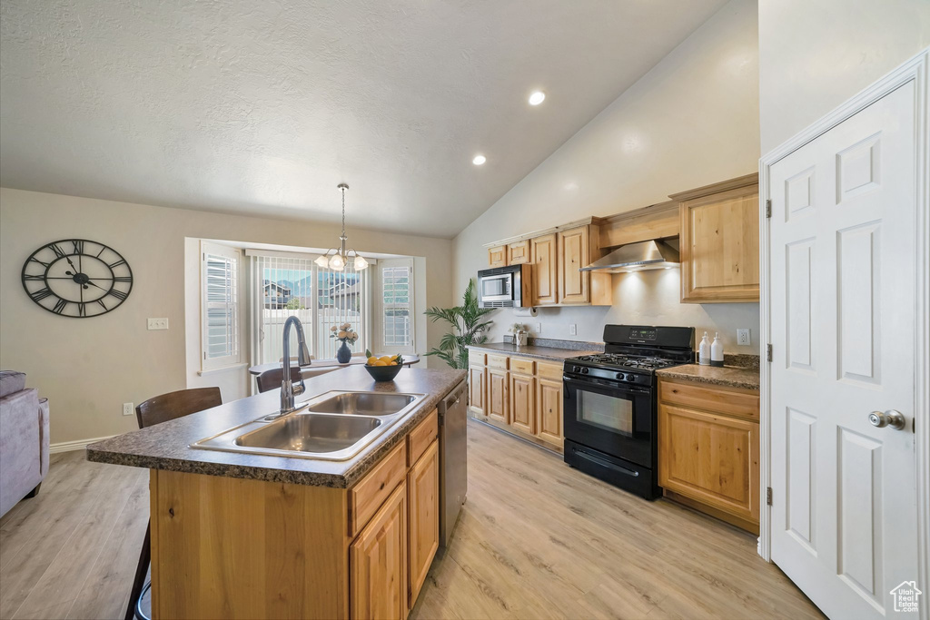 Kitchen featuring appliances with stainless steel finishes, wall chimney range hood, a kitchen island with sink, dark countertops, and light wood-style flooring