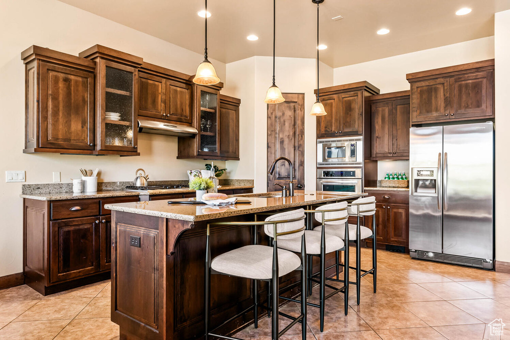 Kitchen featuring appliances with stainless steel finishes, a kitchen island with sink, a breakfast bar area, light tile patterned flooring, and dark brown cabinets