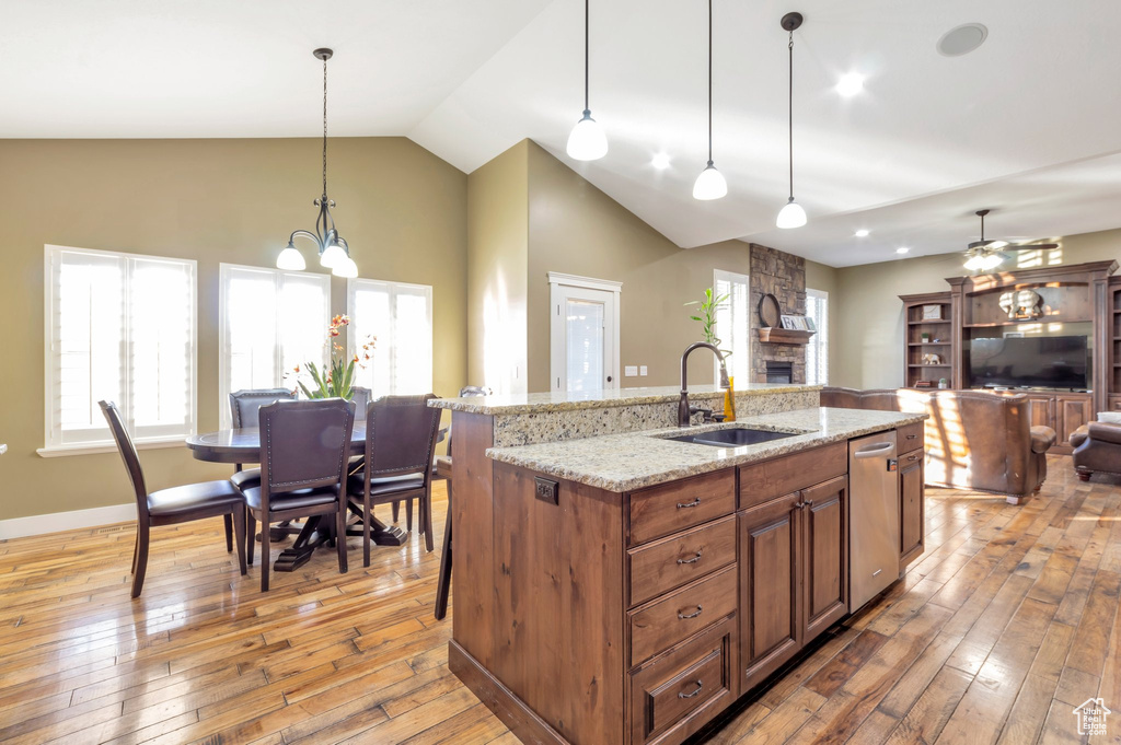 Kitchen featuring a stone fireplace, stainless steel dishwasher, light wood-type flooring, vaulted ceiling, and brown cabinetry
