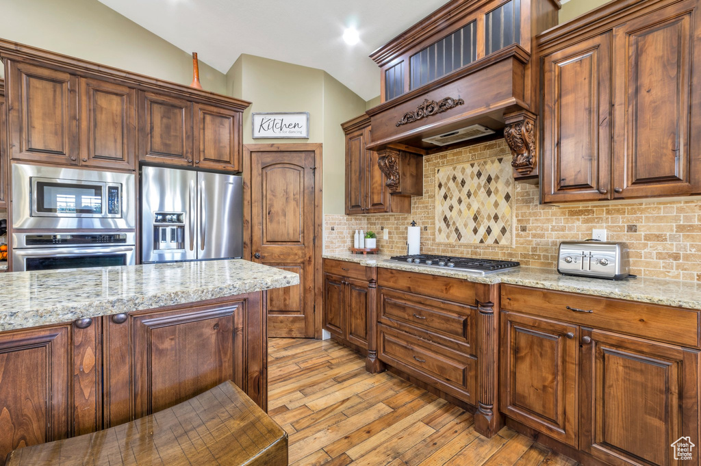 Kitchen featuring appliances with stainless steel finishes, vaulted ceiling, light wood-style floors, decorative backsplash, and light stone countertops