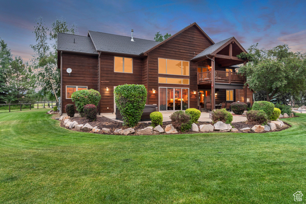 Back of property at dusk featuring a patio area, a lawn, a balcony, and roof with shingles
