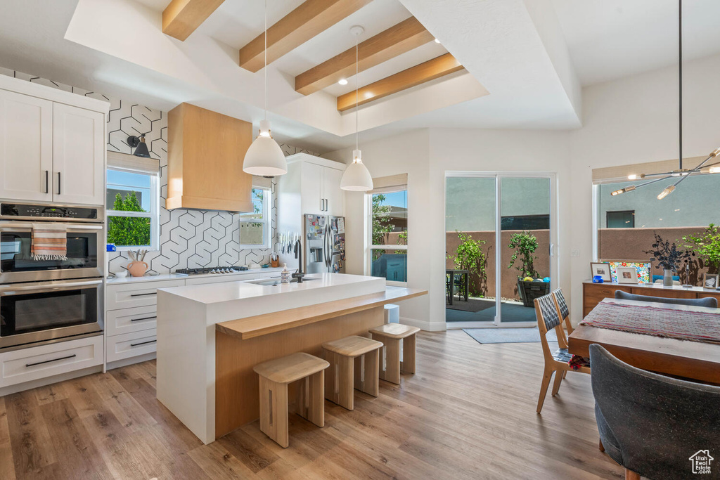 Kitchen featuring stainless steel appliances, decorative backsplash, light countertops, an island with sink, and beamed ceiling