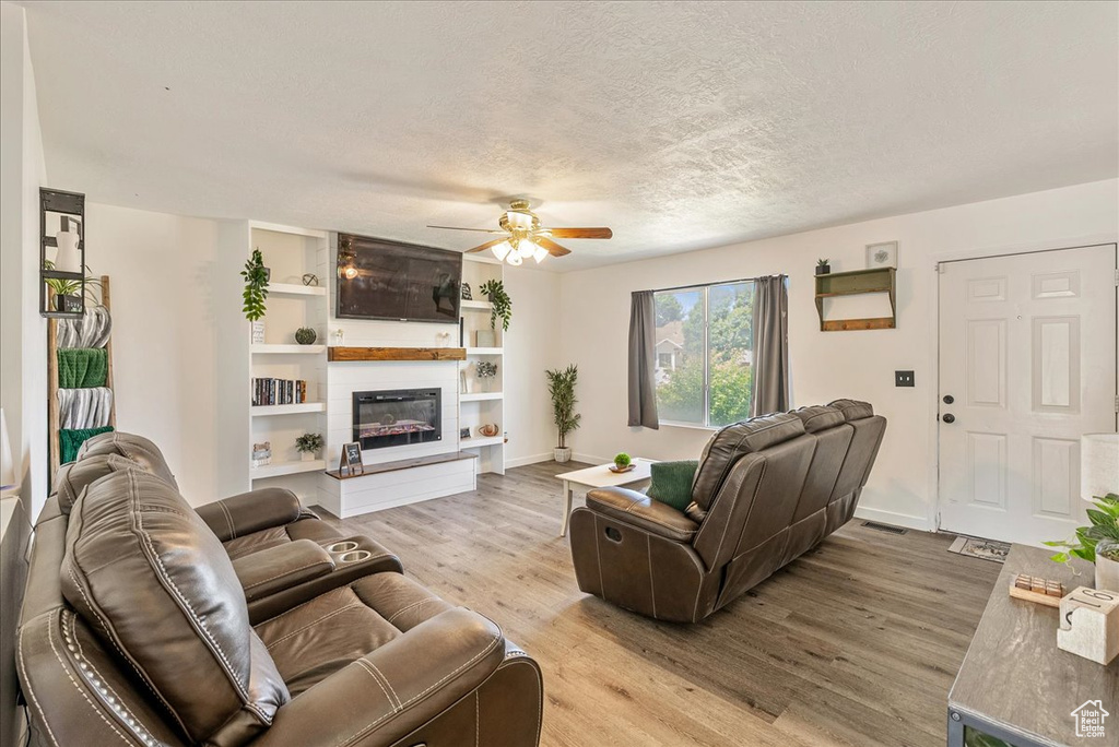Living room featuring light wood-type flooring, a ceiling fan, a textured ceiling, and a glass covered fireplace