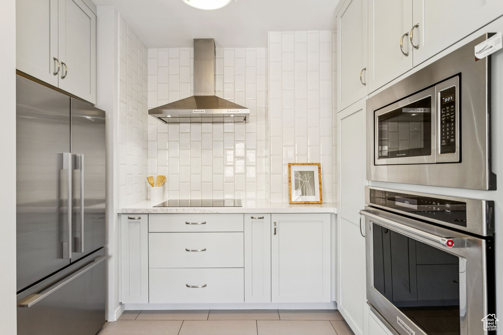 Kitchen featuring appliances with stainless steel finishes, wall chimney exhaust hood, tasteful backsplash, light tile patterned floors, and light stone counters