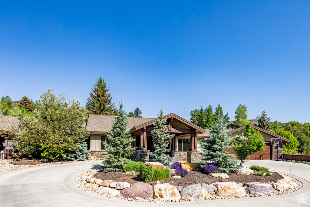 View of property hidden behind natural elements with curved driveway, stone siding, covered porch, a shingled roof, and a garage