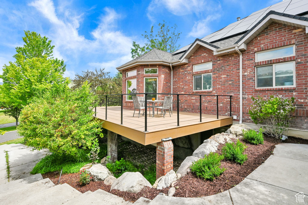 View of side of property with brick siding, a wooden deck, solar panels, outdoor dining area, and a shingled roof