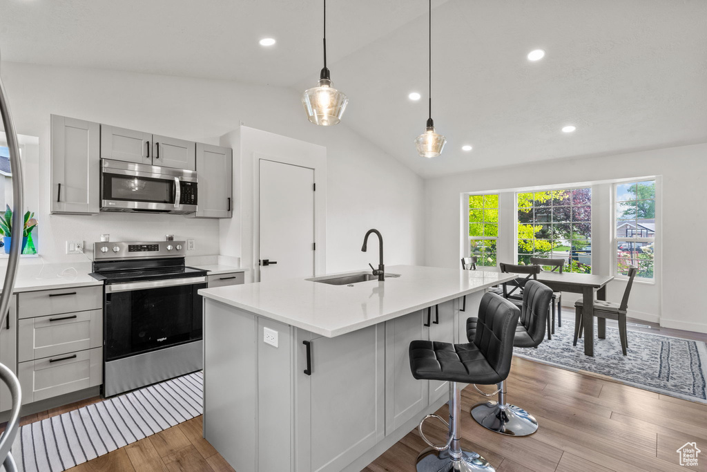 Kitchen with stainless steel appliances, wood finished floors, a center island with sink, light countertops, and vaulted ceiling