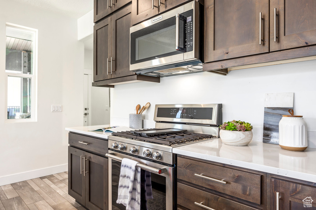 Kitchen featuring stainless steel appliances, dark brown cabinetry, light wood-style flooring, and light stone counters