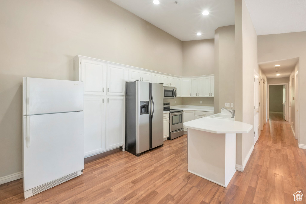 Kitchen featuring appliances with stainless steel finishes, a towering ceiling, a peninsula, white cabinets, and light wood finished floors
