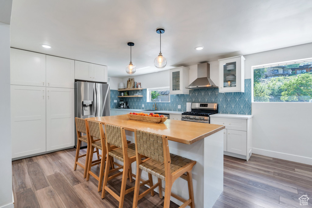 Kitchen featuring wall chimney exhaust hood, stainless steel appliances, decorative backsplash, a breakfast bar, and white cabinetry