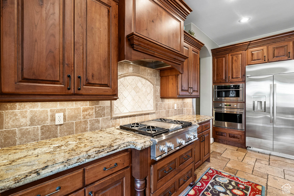Kitchen with appliances with stainless steel finishes, stone tile floors, custom exhaust hood, light stone counters, and tasteful backsplash