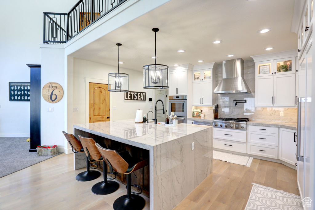 Kitchen with wall chimney range hood, light wood-type flooring, a center island with sink, decorative backsplash, and recessed lighting