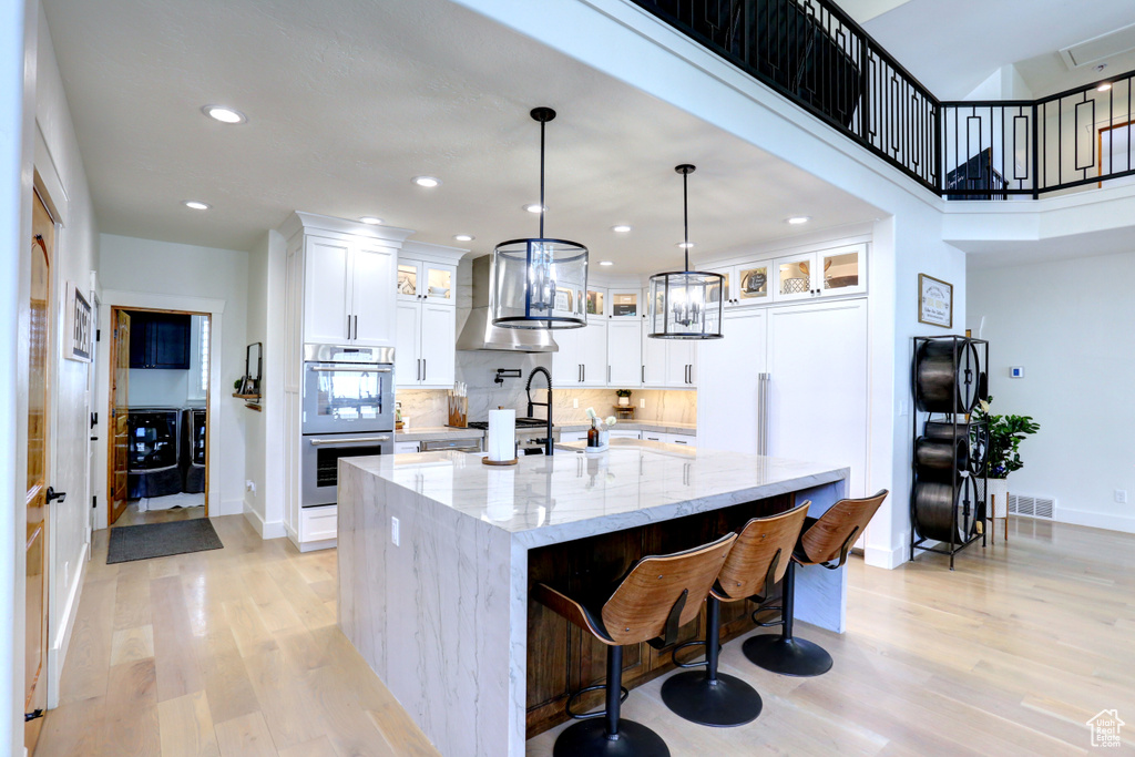 Kitchen with double oven, range hood, light wood-style flooring, a large island with sink, and recessed lighting