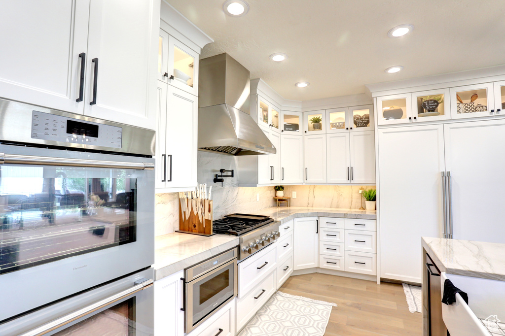 Kitchen with wall chimney exhaust hood, built in appliances, light wood-style flooring, white cabinets, and tasteful backsplash