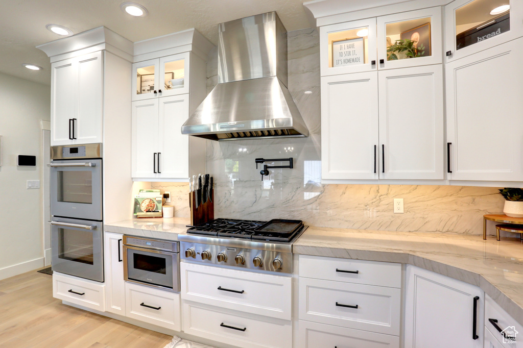 Kitchen with stainless steel appliances, wall chimney range hood, decorative backsplash, light wood-type flooring, and white cabinets
