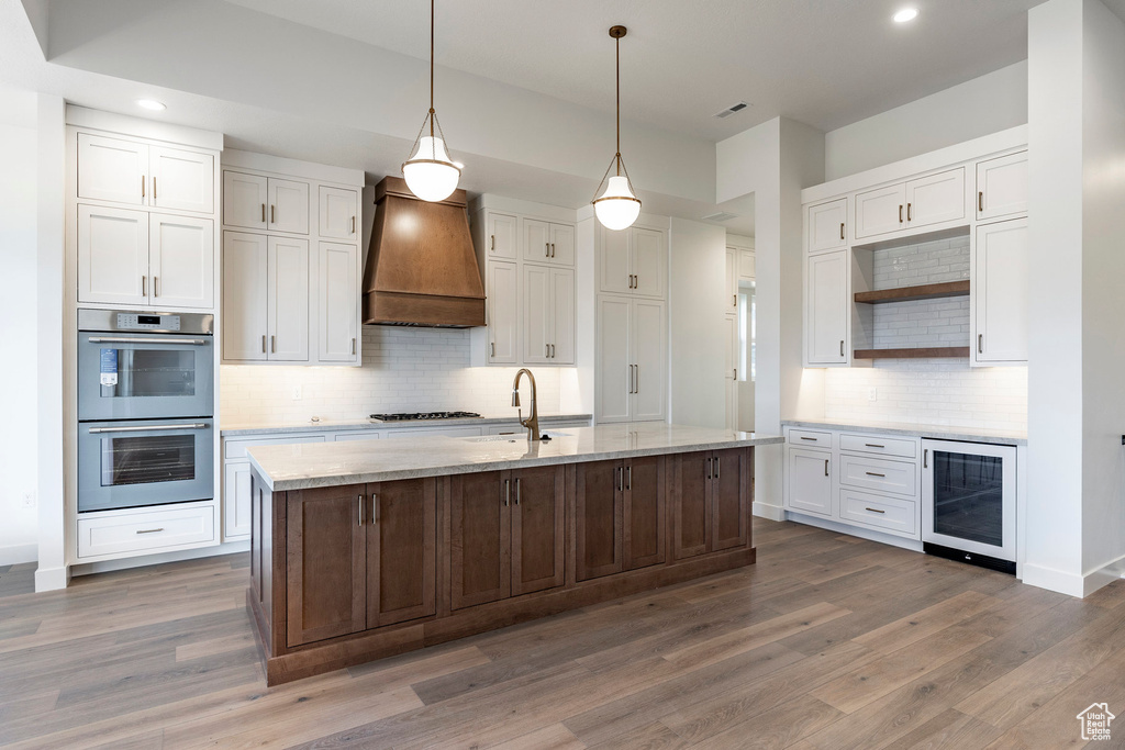 Kitchen featuring wine cooler, custom exhaust hood, stainless steel double oven, open shelves, and white cabinetry