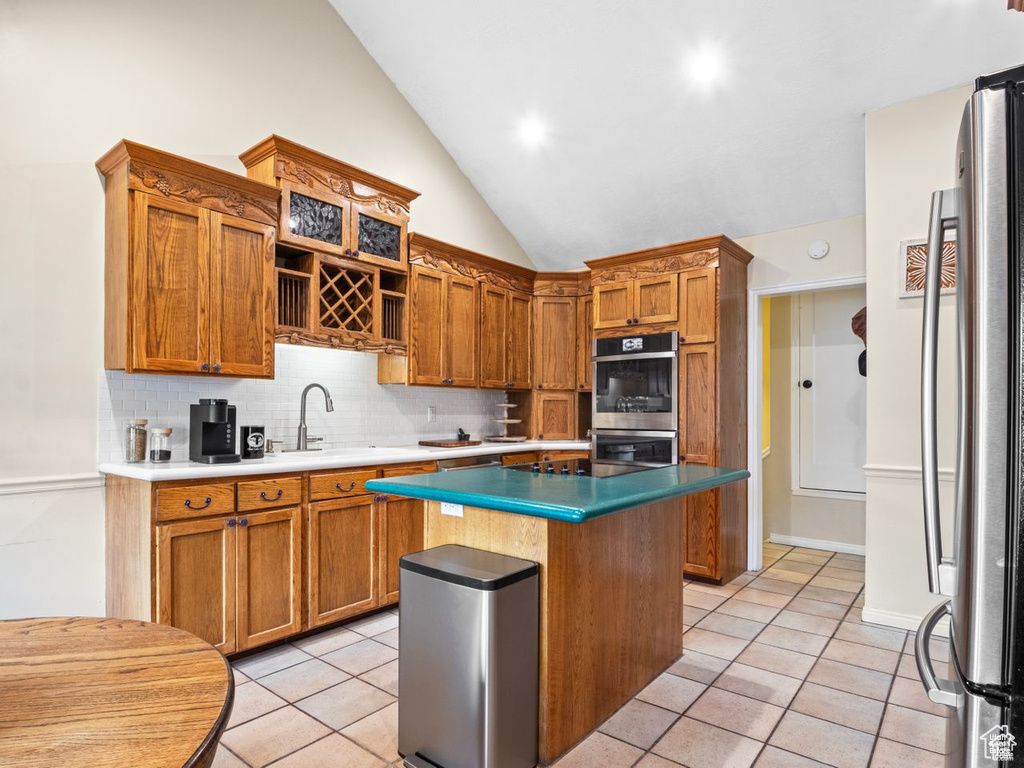 Kitchen featuring appliances with stainless steel finishes, light tile patterned flooring, decorative backsplash, a kitchen island, and brown cabinetry