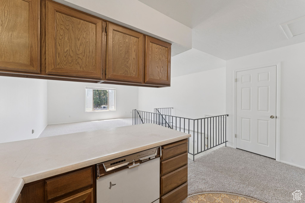 Kitchen featuring white dishwasher, light colored carpet, brown cabinetry, and light countertops