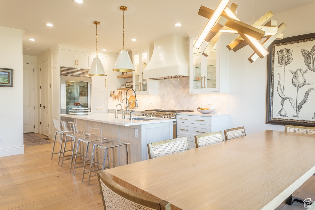 Kitchen featuring custom range hood, light wood-style flooring, an island with sink, tasteful backsplash, and a breakfast bar