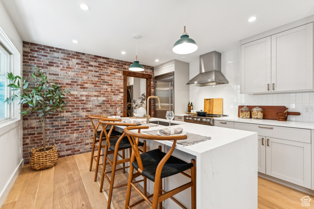 Kitchen with wall chimney exhaust hood, built in fridge, brick wall, light wood finished floors, and a center island with sink