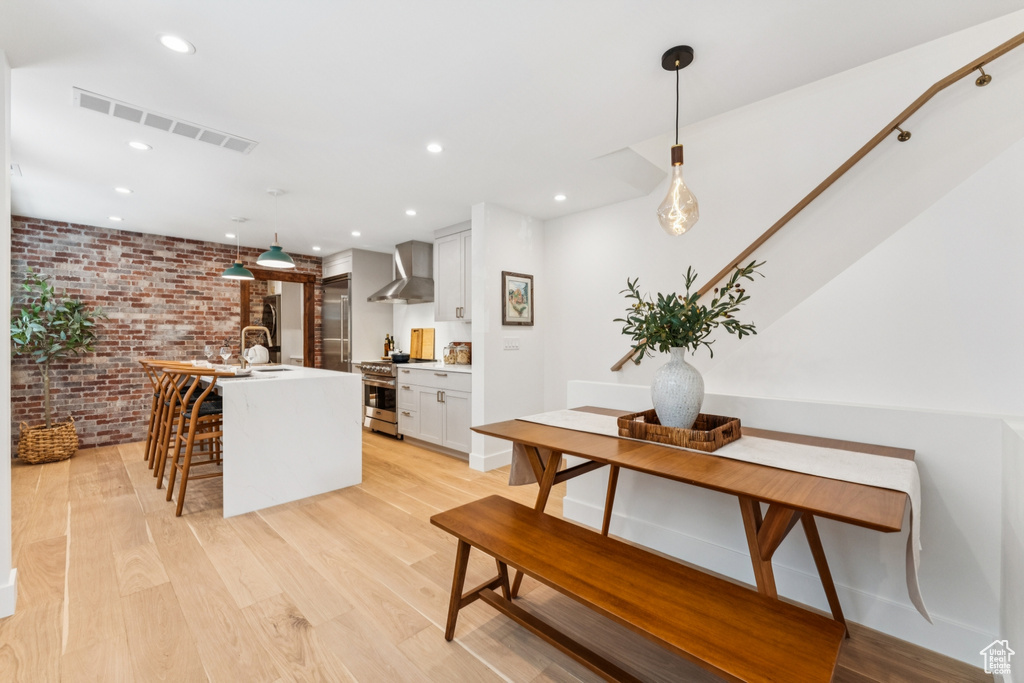 Kitchen with brick wall, wall chimney range hood, stainless steel stove, a kitchen island with sink, and light wood finished floors
