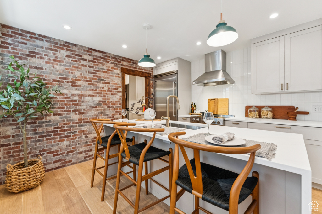 Kitchen featuring wall chimney exhaust hood, stainless steel built in fridge, brick wall, light wood-type flooring, and a breakfast bar area