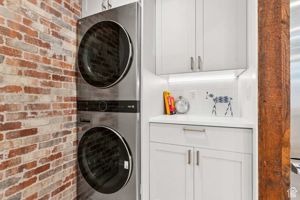 Washroom featuring stacked washing machine and dryer, brick wall, and cabinet space