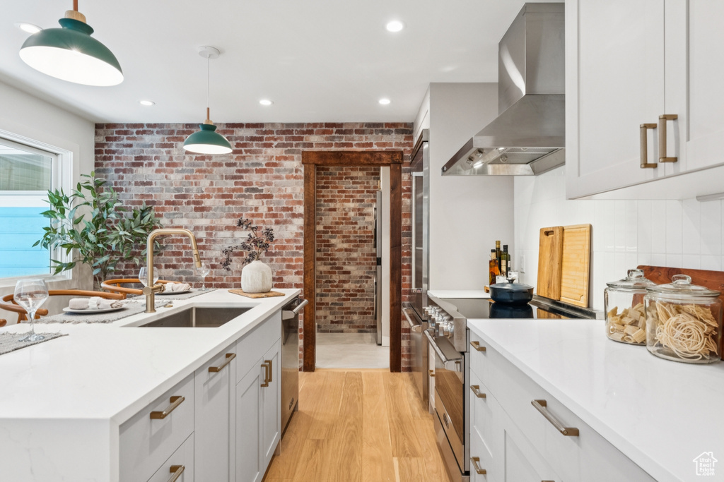 Kitchen with wall chimney exhaust hood, appliances with stainless steel finishes, brick wall, light countertops, and light wood-style flooring