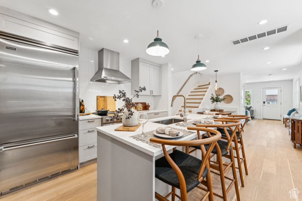 Kitchen featuring built in fridge, wall chimney exhaust hood, light wood-style floors, backsplash, and a kitchen bar