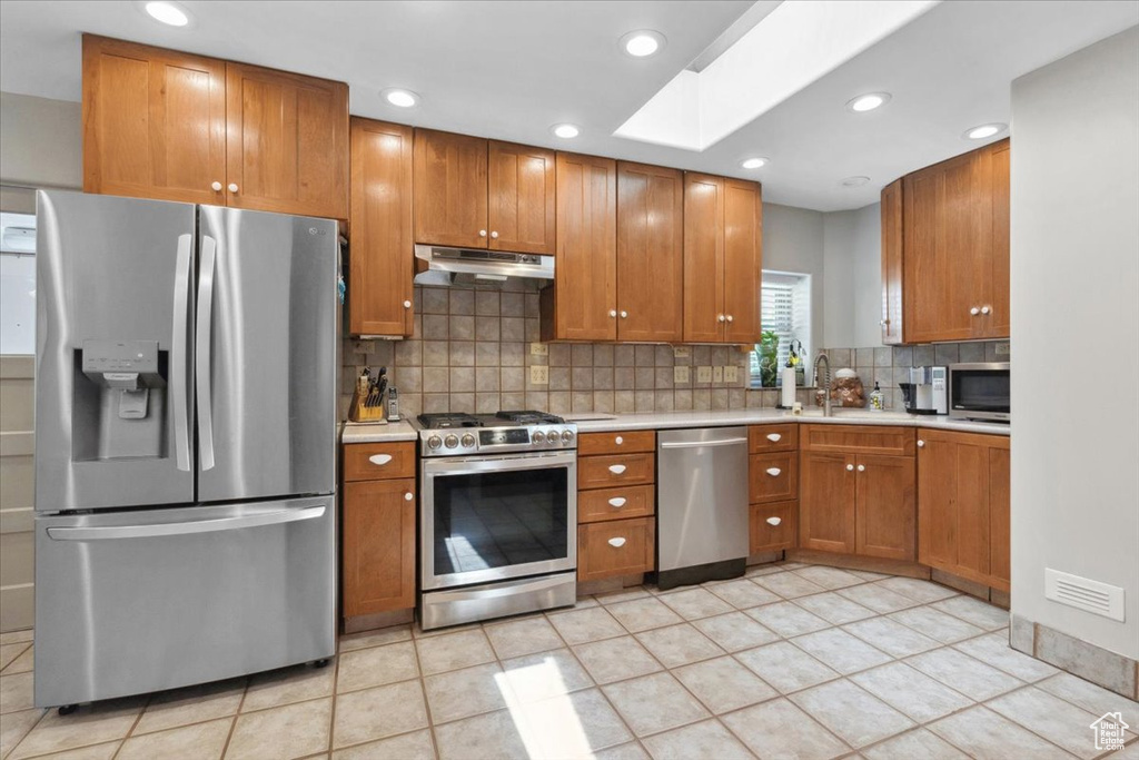 Kitchen featuring stainless steel appliances, under cabinet range hood, light countertops, tasteful backsplash, and brown cabinets