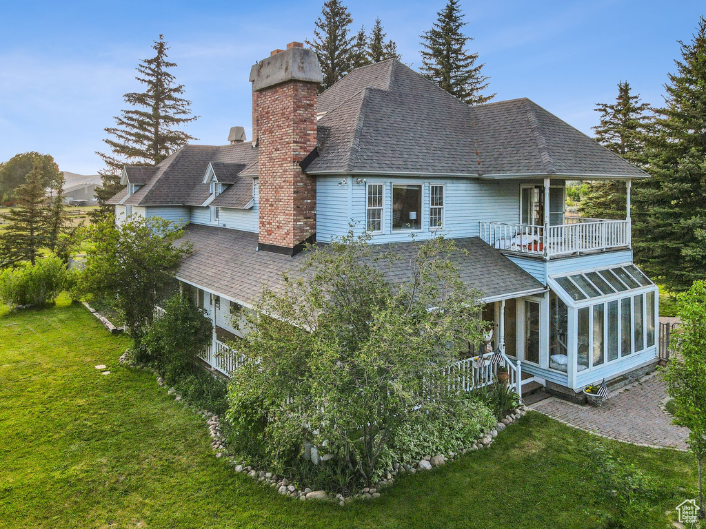 Rear view of property featuring a chimney, a balcony, a sunroom, a shingled roof, and a lawn