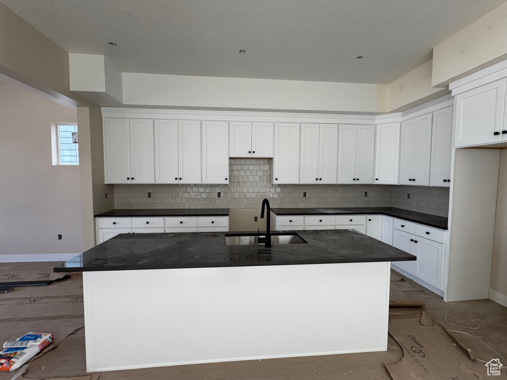 Kitchen featuring dark countertops, decorative backsplash, white cabinets, and an island with sink