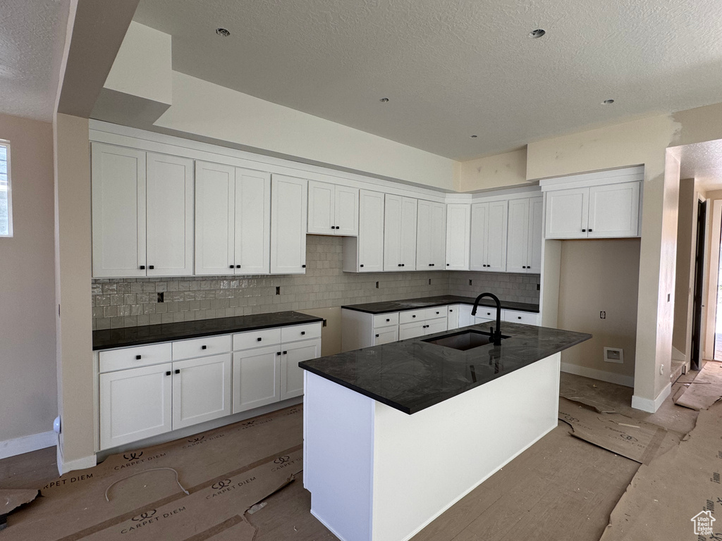 Kitchen featuring a textured ceiling, decorative backsplash, white cabinetry, dark countertops, and an island with sink