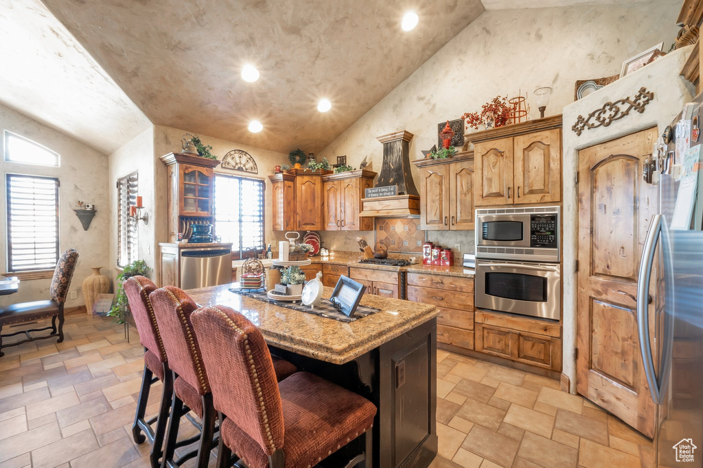 Kitchen with vaulted ceiling, stainless steel appliances, custom exhaust hood, light stone finish flooring, and light stone countertops