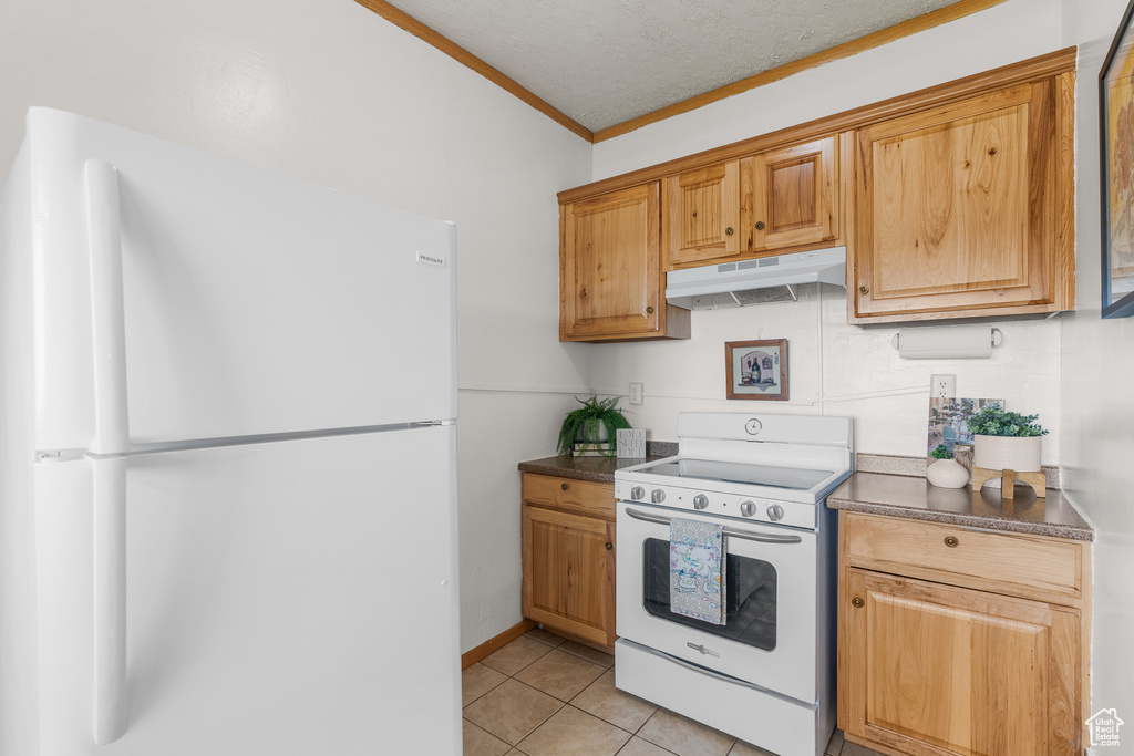 Kitchen with white appliances, under cabinet range hood, crown molding, dark countertops, and light tile patterned floors
