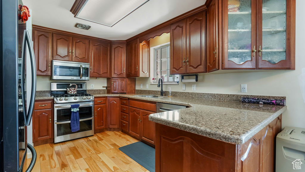 Kitchen featuring appliances with stainless steel finishes, light wood finished floors, glass insert cabinets, a peninsula, and dark stone counters