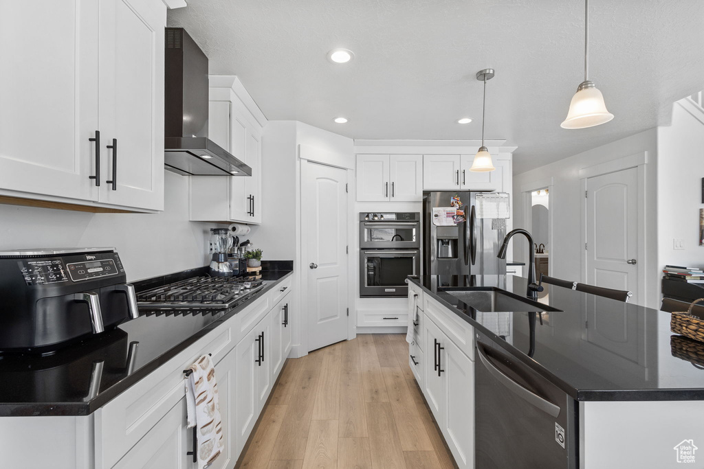 Kitchen featuring wall chimney range hood, stainless steel appliances, dark countertops, a kitchen island with sink, and light wood finished floors