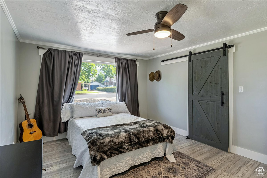 Bedroom with ornamental molding, a textured ceiling, a barn door, light wood-style flooring, and ceiling fan