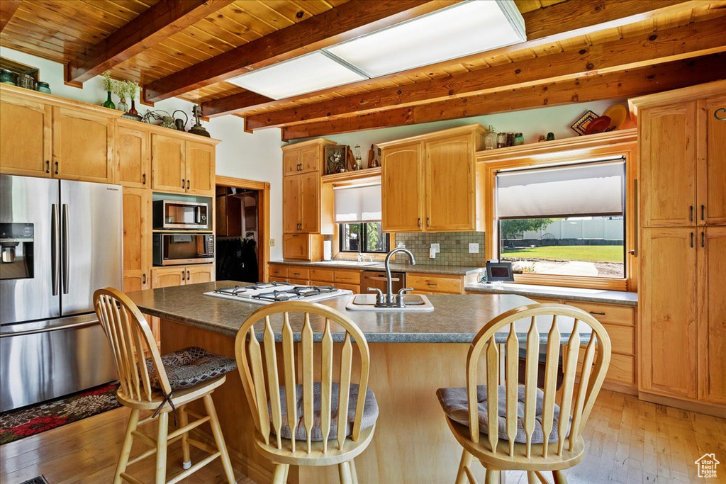Kitchen with appliances with stainless steel finishes, light wood-style floors, a wooden ceiling with exposed beams, decorative backsplash, and an island with sink