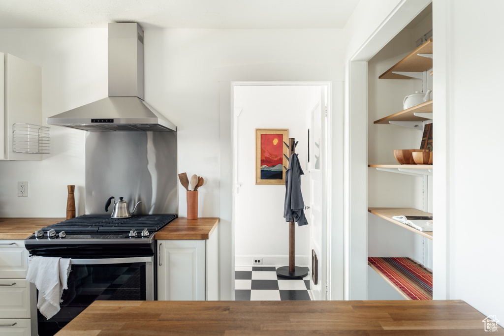 Kitchen with wall chimney exhaust hood, stainless steel gas range oven, wooden counters, tile patterned floors, and white cabinetry