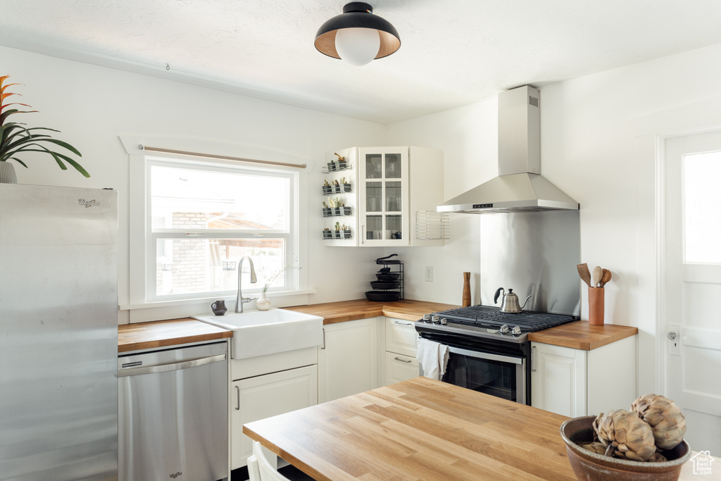 Kitchen with butcher block countertops, wall chimney range hood, appliances with stainless steel finishes, glass insert cabinets, and white cabinetry