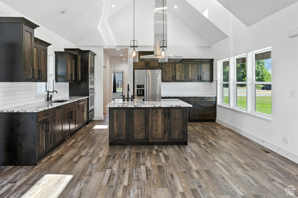 Kitchen featuring stainless steel appliances, dark brown cabinetry, decorative backsplash, a center island with sink, and high vaulted ceiling
