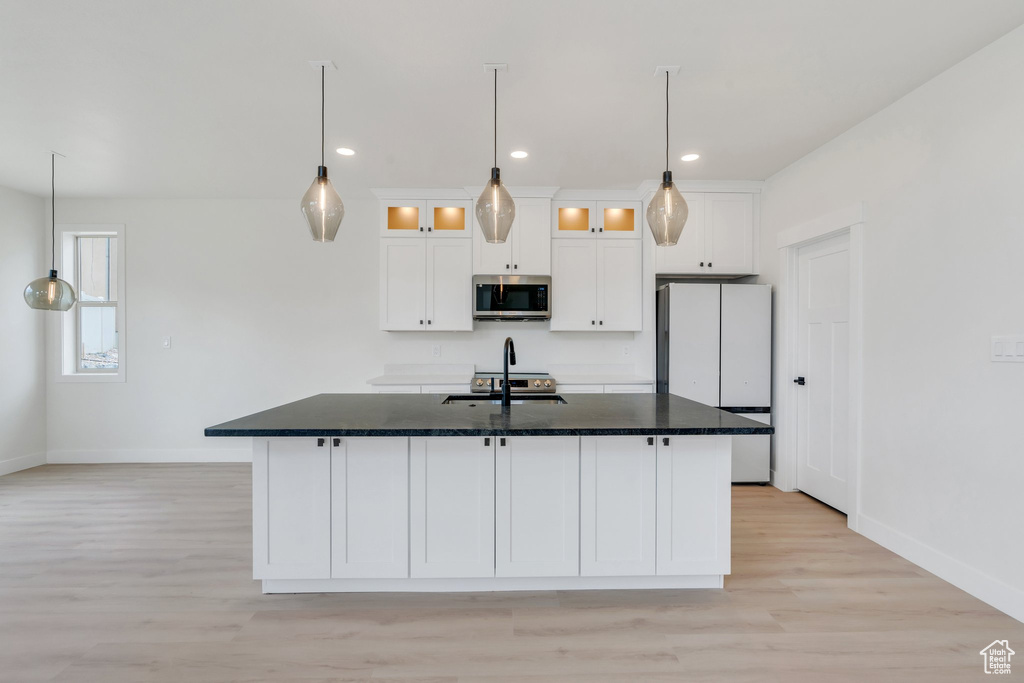 Kitchen featuring stainless steel microwave, freestanding refrigerator, white cabinetry, a kitchen island with sink, and light wood-style floors