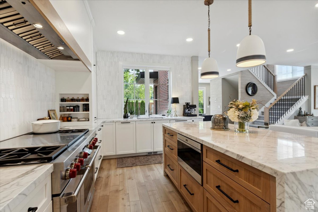Kitchen with appliances with stainless steel finishes, wall chimney exhaust hood, light wood-type flooring, light stone counters, and white cabinetry