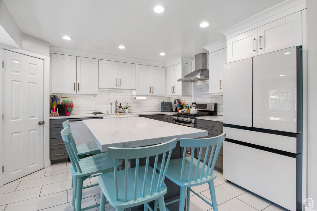 Kitchen featuring freestanding refrigerator, stainless steel range with gas stovetop, wall chimney range hood, light tile patterned flooring, and white cabinetry